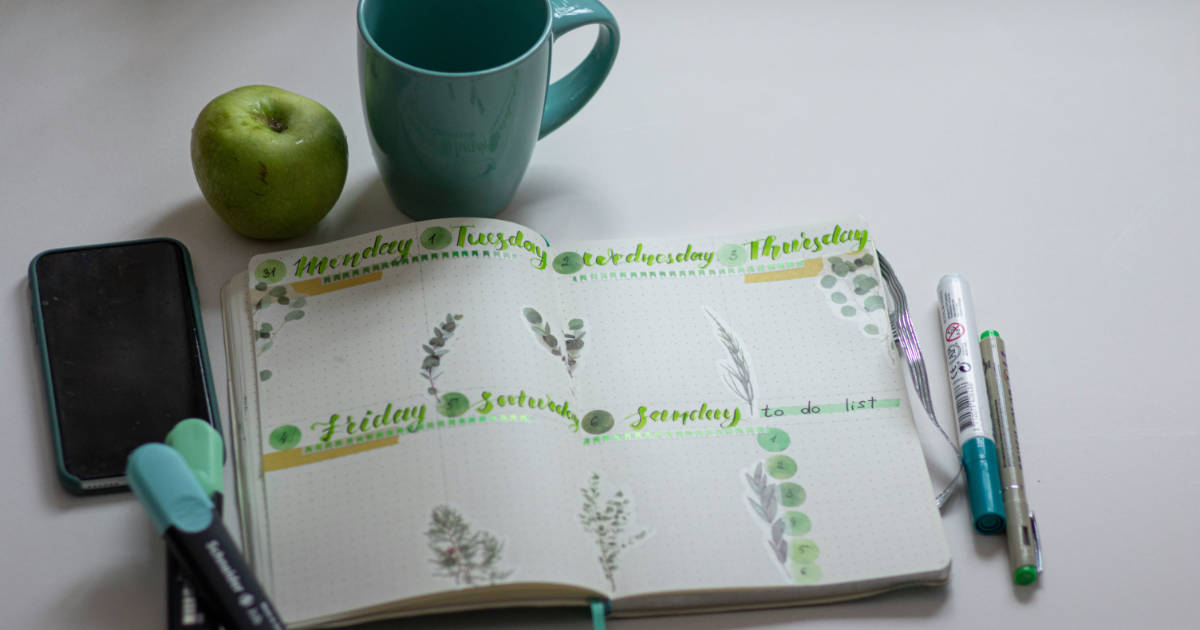 Ein aufgeschlagenes Buch mit einer Wochenübersicht, einer türkisen Keramiktasse, Stifen und einem grünen Apfel daneben. (Foto: Elena Mozhvilo)