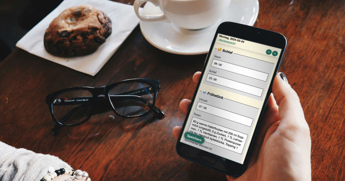 Nahaufnahme auf ein Smartphone das eine Person in der Hand hält. Das Smartphone zeigt die Nutrition Logs Protokoll-Ansicht. Im Hintergurnd auf braunem Holztisch eine Tasse Kaffee, ein Keks auf einer weißen Serviette und eine schwarze Brille. (Foto: Patrick Tomasso)